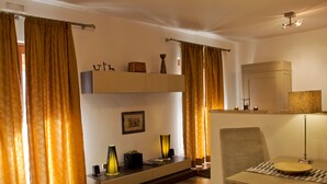 A modern living room with a sofa, a dining table, and a TV. There are curtains, a lamp, and a shelf with decorative items. - Ecolodge Selvitelle - Escape the hectic and submerge in the nature of Cilento (Cicerale)