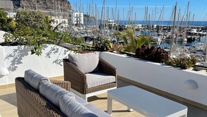 Een terras met rieten meubels, een witte tafel en uitzicht op een jachthaven met boten.
