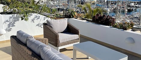 A terrace with wicker furniture, a white table, and a view of a marina with boats.