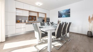 Eine moderne Küche mit weißen Schränken, ein Essbereich mit einem weißen Tisch und grauen Stühlen sowie ein Gemälde einer Berglandschaft an der Wand.