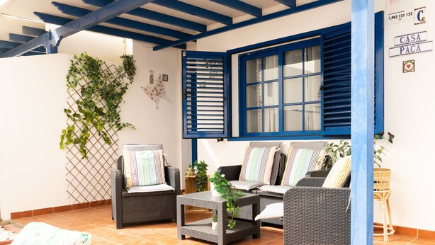 Een gezellig terras met rieten meubels, een betegelde vloer en een blauw raamkozijn.