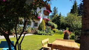 Ein Garten mit einer Holzbank und einem Tisch, umgeben von üppigem Grün und einem Gebäude mit Balkonen.