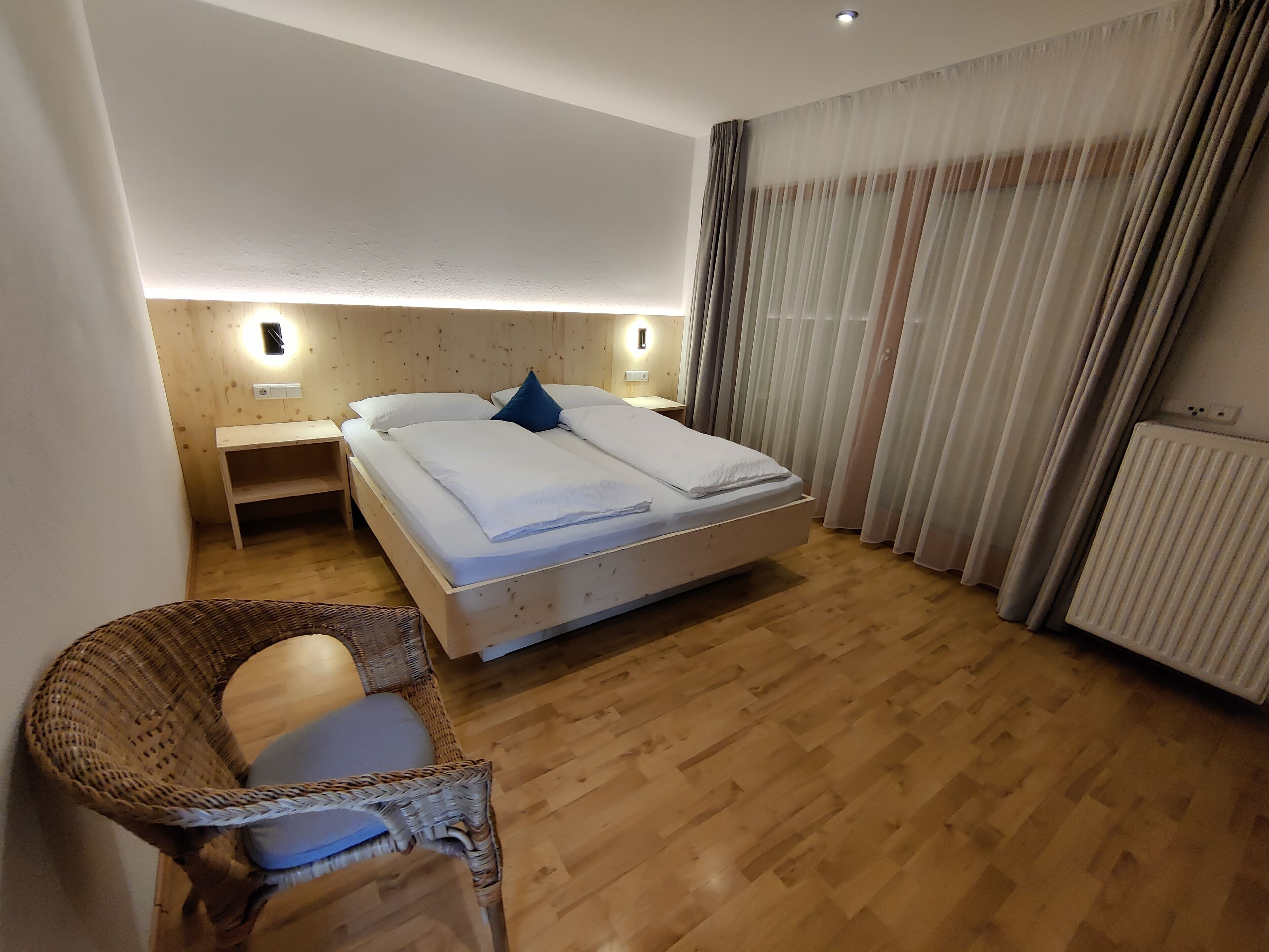 Une chambre d’hôtel avec un plancher en bois, un lit avec une literie blanche, une chaise en osier, une table de chevet et un luminaire fixé au mur.