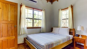 Ein hölzernes Bett mit karierter Bettdecke, zwei Fenster mit Vorhängen, ein Nachttisch aus Holz mit einer Lampe und eine Holztür.