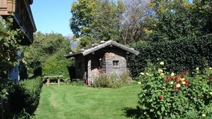 Eine rustikale Holzhütte mit geneigtem Dach, umgeben von einem gepflegten Garten mit verschiedenen Pflanzen und Blumen.