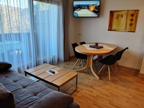 Un salon comprenant un canapé, une table basse, une table à manger avec des chaises, un téléviseur fixé au mur et une fenêtre ornée de rideaux légers.