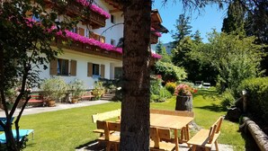 A building with a balcony adorned with pink flowers, a wooden table and benches in the foreground, and a well-maintained garden with various plants and a large tree. - Residence Klementhof #4 (Natz)