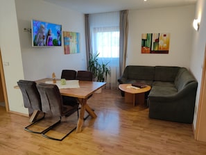 Ett vardagsrum med ett matbord i trä, två stolar, en grön soffa och en tv som är monterad på väggen.
