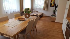 A dining area with a wooden table, chairs, a vase with flowers, and a sofa in the background.