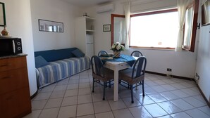 Un espacio habitable compacto con kitchenette, una zona de comedor con mesa y sillas, un sofá y un amplio ventanal con cortinas.