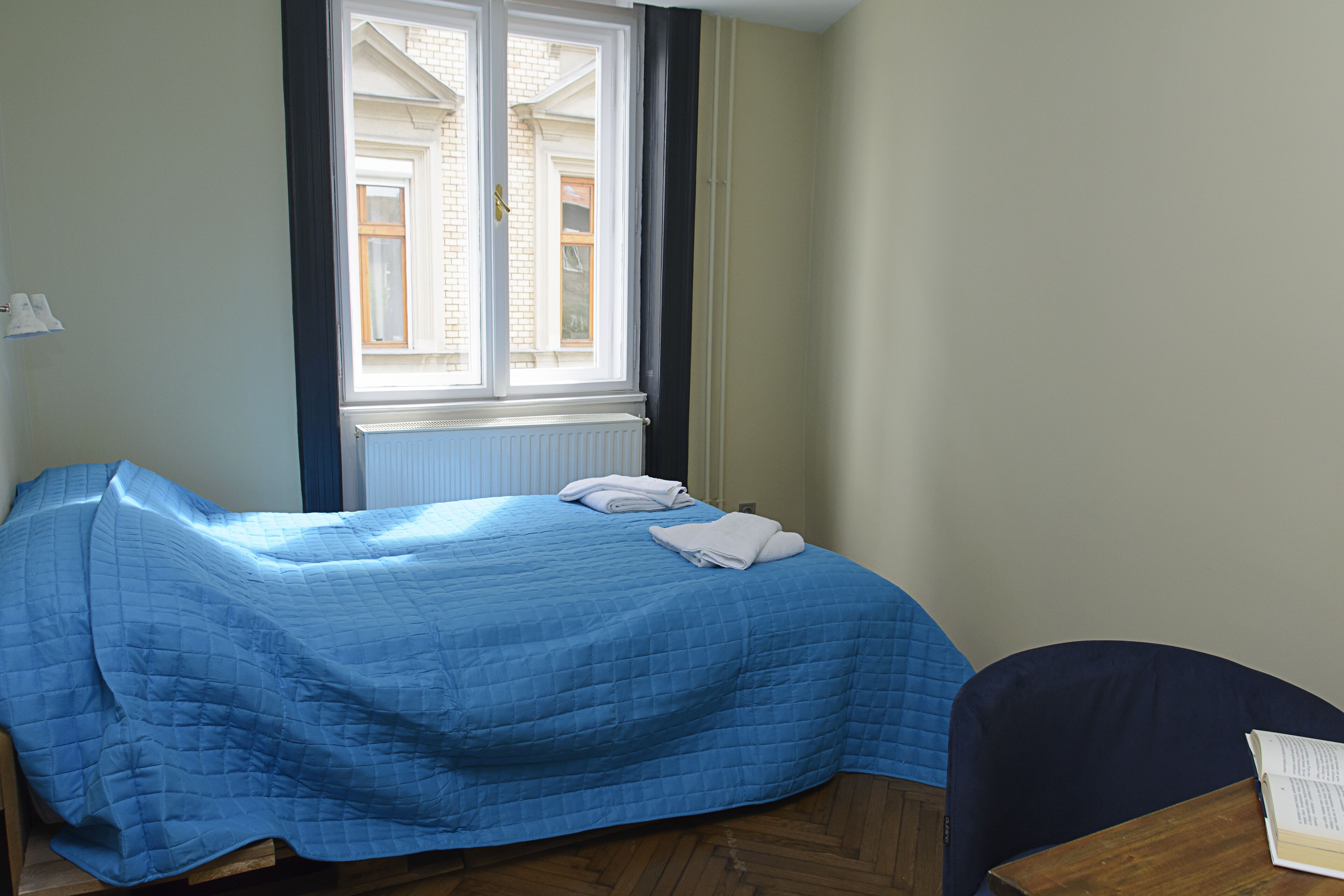 A bedroom with a bed covered in blue bedding, a window with blinds, a radiator, and a chair with a book.