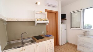 Une cuisine équipée d’un évier, de placards, d’un plan de travail, d’une porte en bois, d’une petite étagère avec des objets décoratifs, d’une télévision et d’une fenêtre.