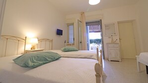 Un dormitorio con dos camas, una mesita de noche con lámpara, un televisor montado en la pared y una ventana con cortinas.