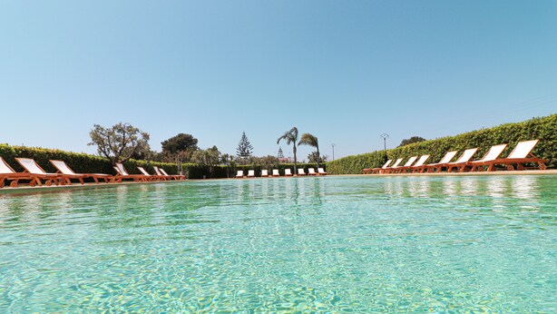 Una piscina cristalina con sillones de descanso a ambos lados, rodeada de setos y árboles perfectamente cuidados.