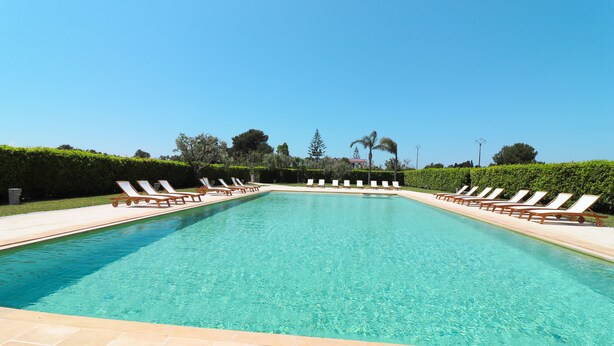 Una piscina rettangolare con acqua cristallina e azzurra, circondata da un solarium piastrellato.