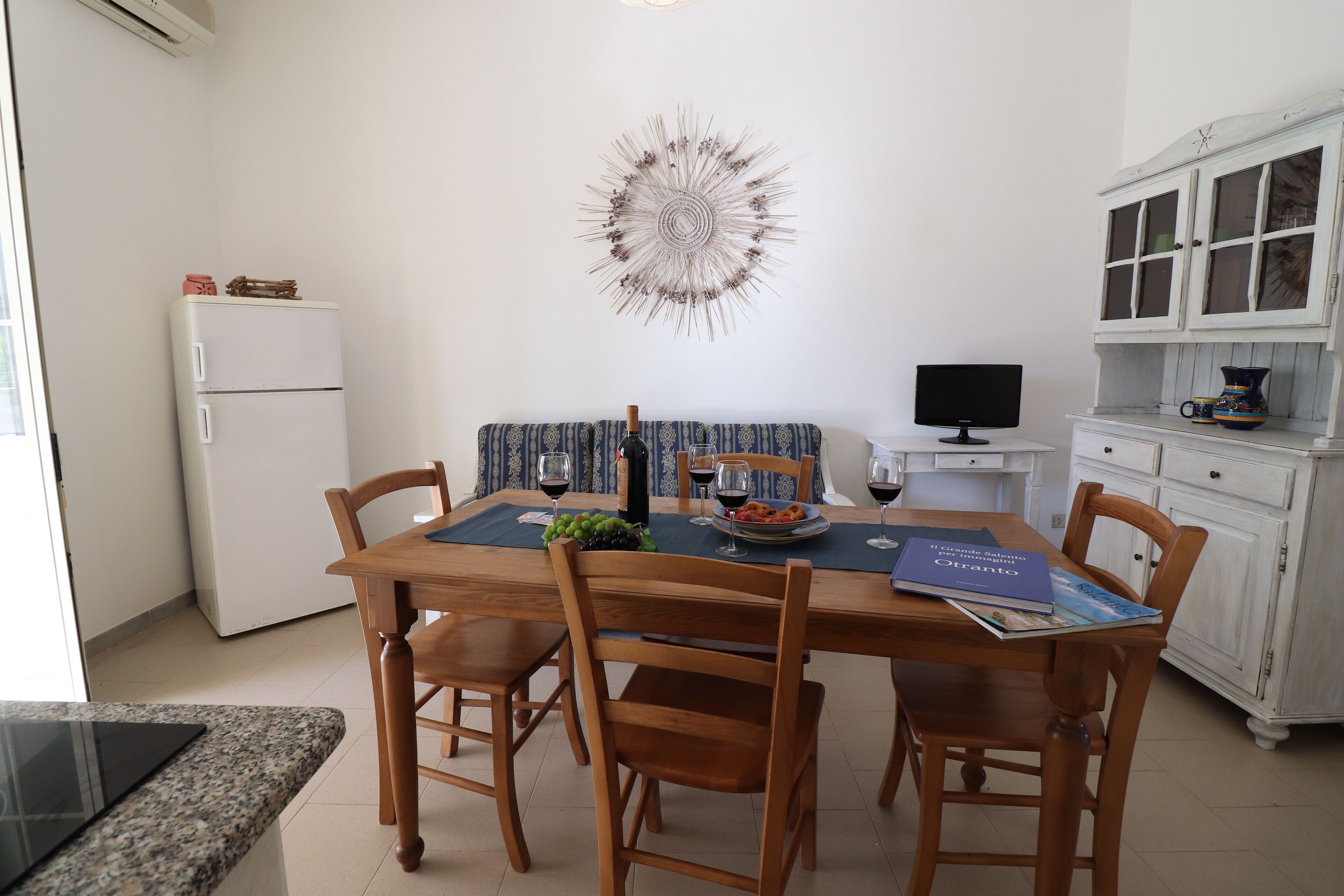 Ein Essbereich mit einem Holztisch, vier Stühlen, einer Wanddekoration, einem weißen Kühlschrank, einem Fernseher und einem Schrank mit Geschirr.