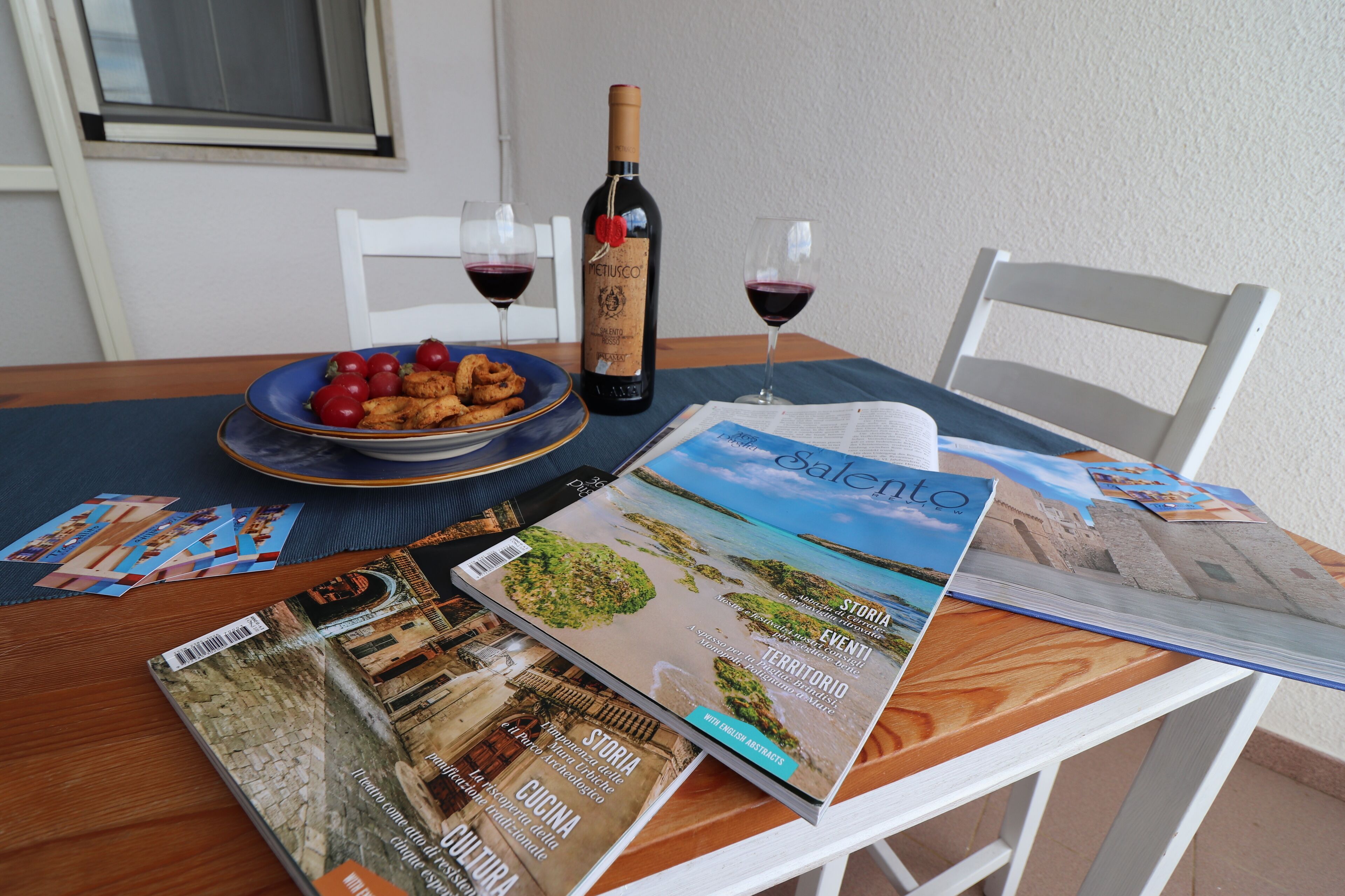 A dining table with a bottle of wine, two glasses of wine, a plate of food, and several magazines.