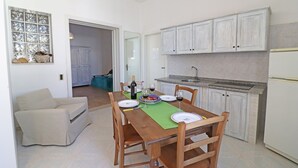 A dining area with a wooden table set for two, a sofa, and a kitchen with white cabinets and a refrigerator.