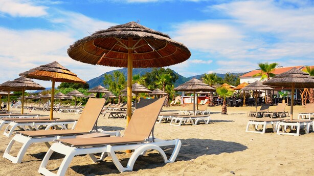 Ein Strand mit weißen Liegestühlen und strohgedeckten Sonnenschirmen, vor einer Kulisse aus Bergen und Palmen.