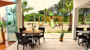 Ein Patio mit Tisch und Stühlen, Blick auf einen Spielplatz und ein Strand im Hintergrund.