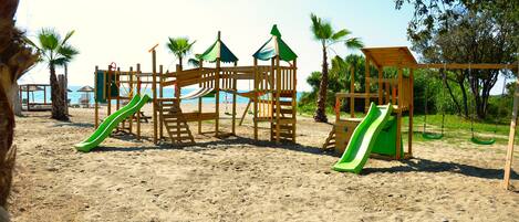 A wooden playground with slides and climbing structures on a sandy beach.
