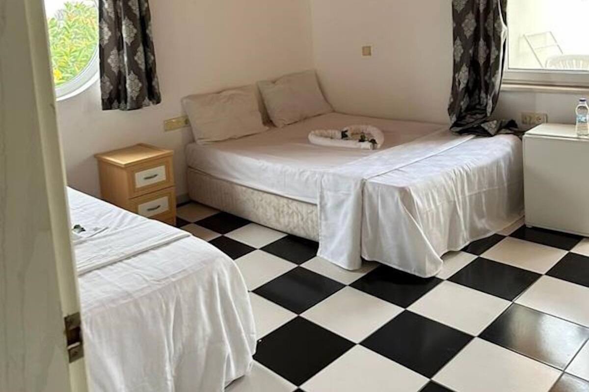 A room with a checkered floor, a bed with a white mattress and matching bedding, a small bedside table with a lamp, and a window with curtains.