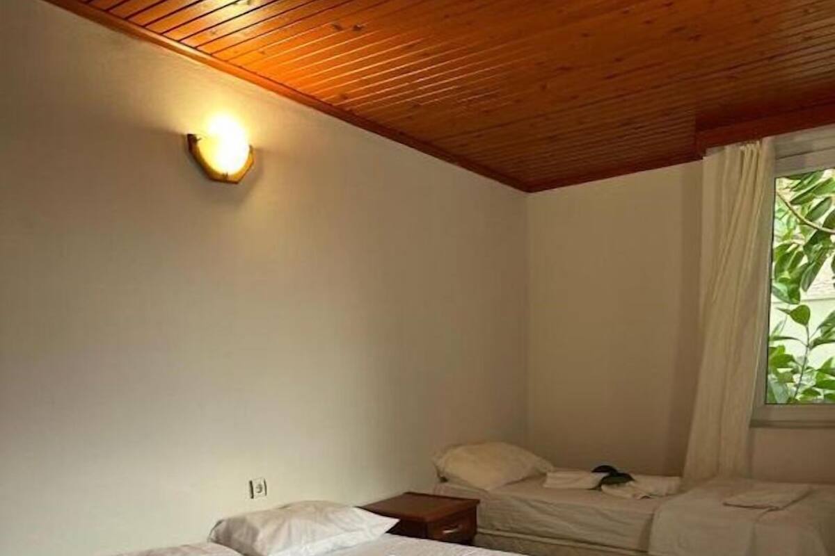 A bedroom with two beds, a wooden ceiling, a nightstand, and a window with curtains.