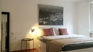 A bedroom with a bed, bedside table, lamp, and a cityscape painting on the wall. - 2 rooms with balcony, central, quiet location (St. Gallen)