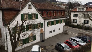 A two-story building with a white facade and green shutters, parked cars in front, and a hilly landscape in the background. - 2 rooms with balcony, central, quiet location (St. Gallen)