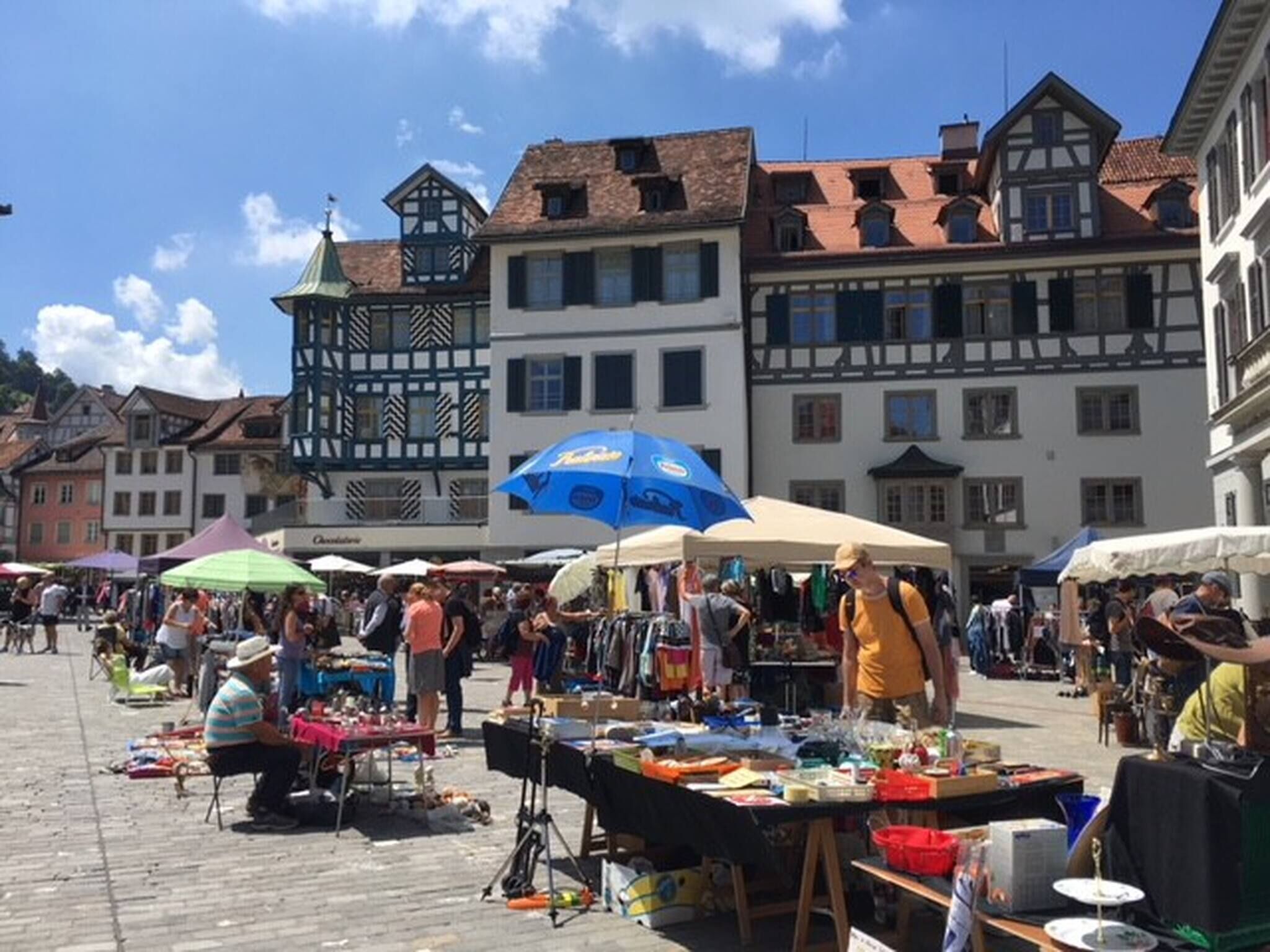 A bustling outdoor market with vendors and visitors, set against a backdrop of traditional European architecture.