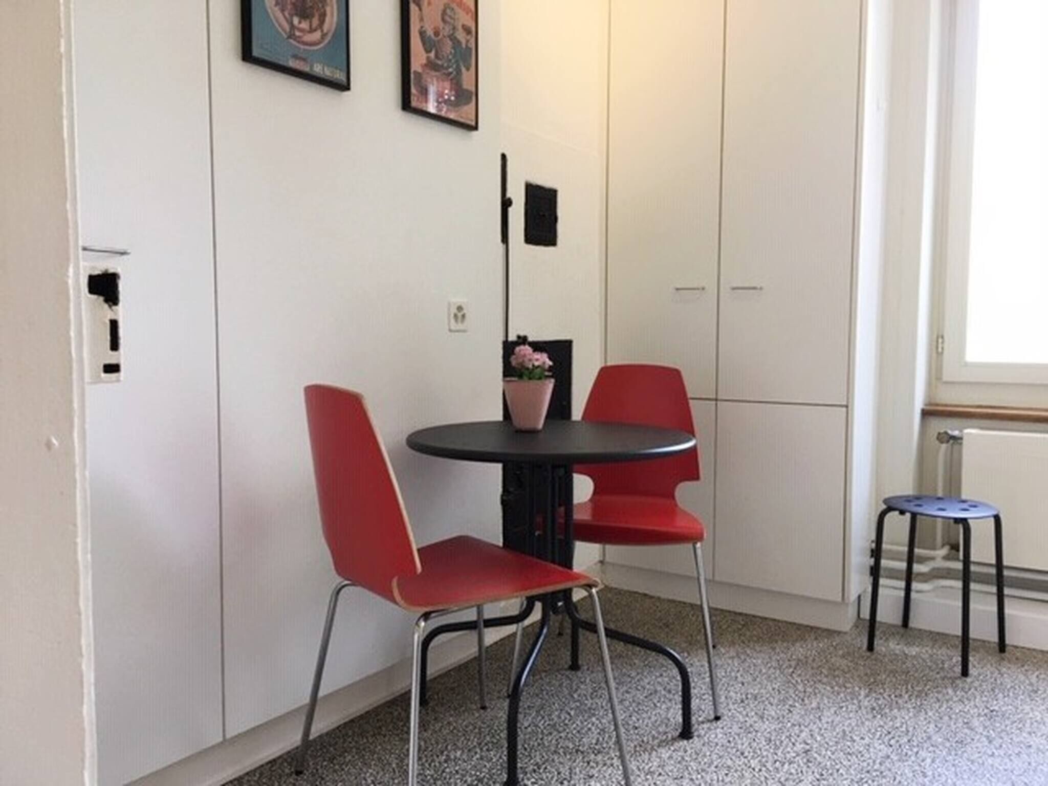Een kleine kamer met een ronde tafel, twee rode stoelen, een kamerplant en een radiator.