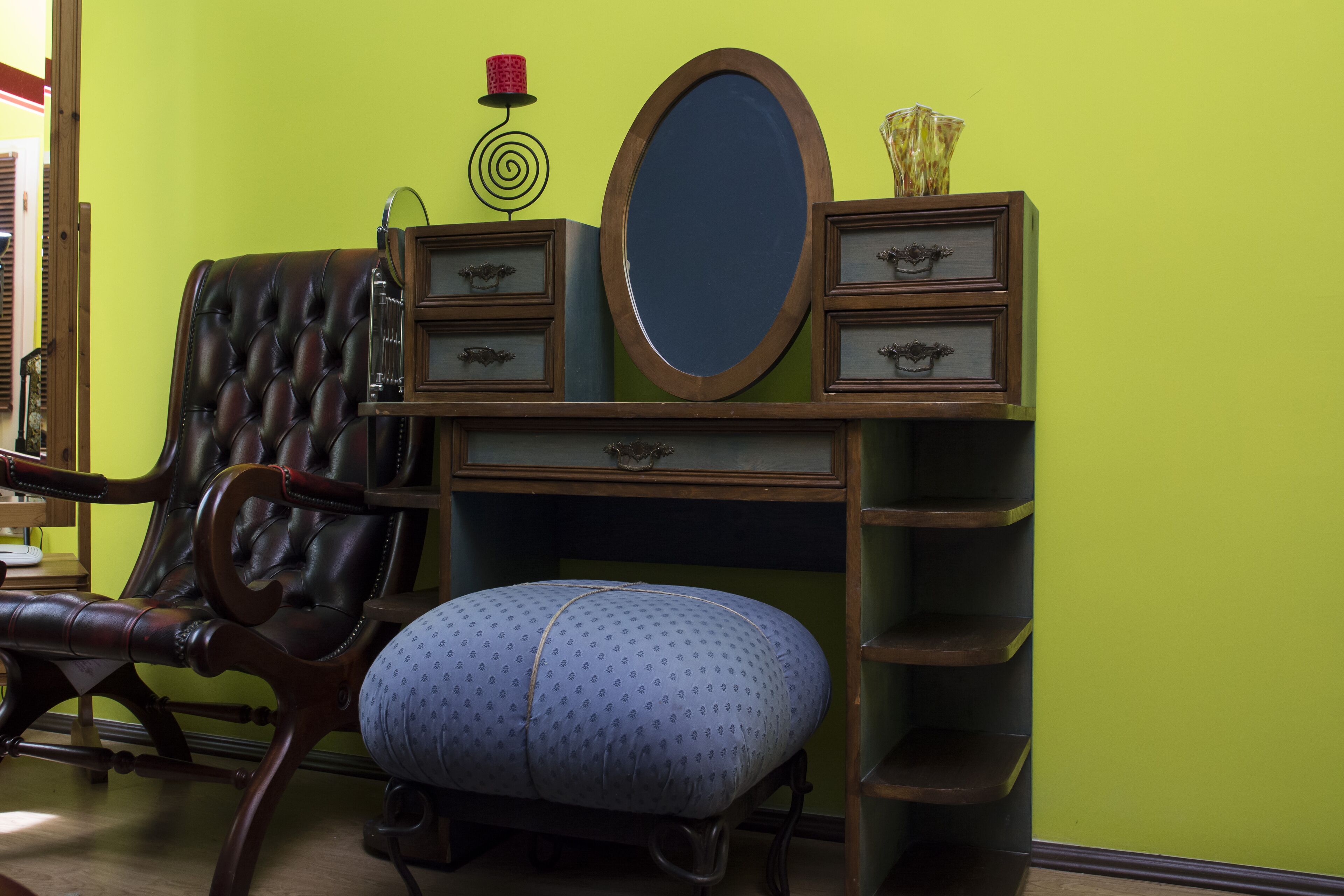 A room with a green wall, a wooden vanity with drawers, a mirror, and a tufted leather chair.
