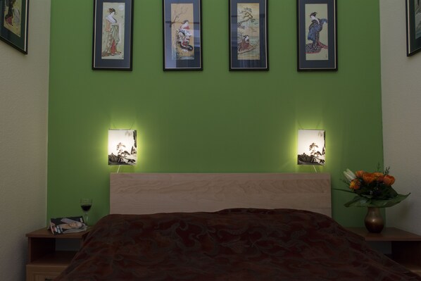 Ein Schlafzimmer mit grüner Wand, einem hölzernen Kopfteil, einem Bett mit gemustertem Überwurf, einem Nachttisch mit einem Buch und einem Weinglas sowie einer Vase mit Blumen.
