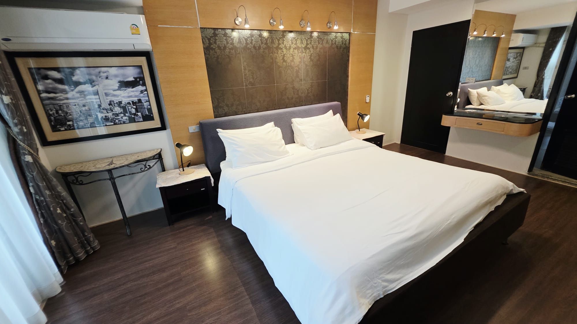 Ein Hotelzimmer mit einem großen Bett, einem Nachttisch, einem Schreibtisch mit Lampe, einem Wand-Fernseher und einem Spiegel.