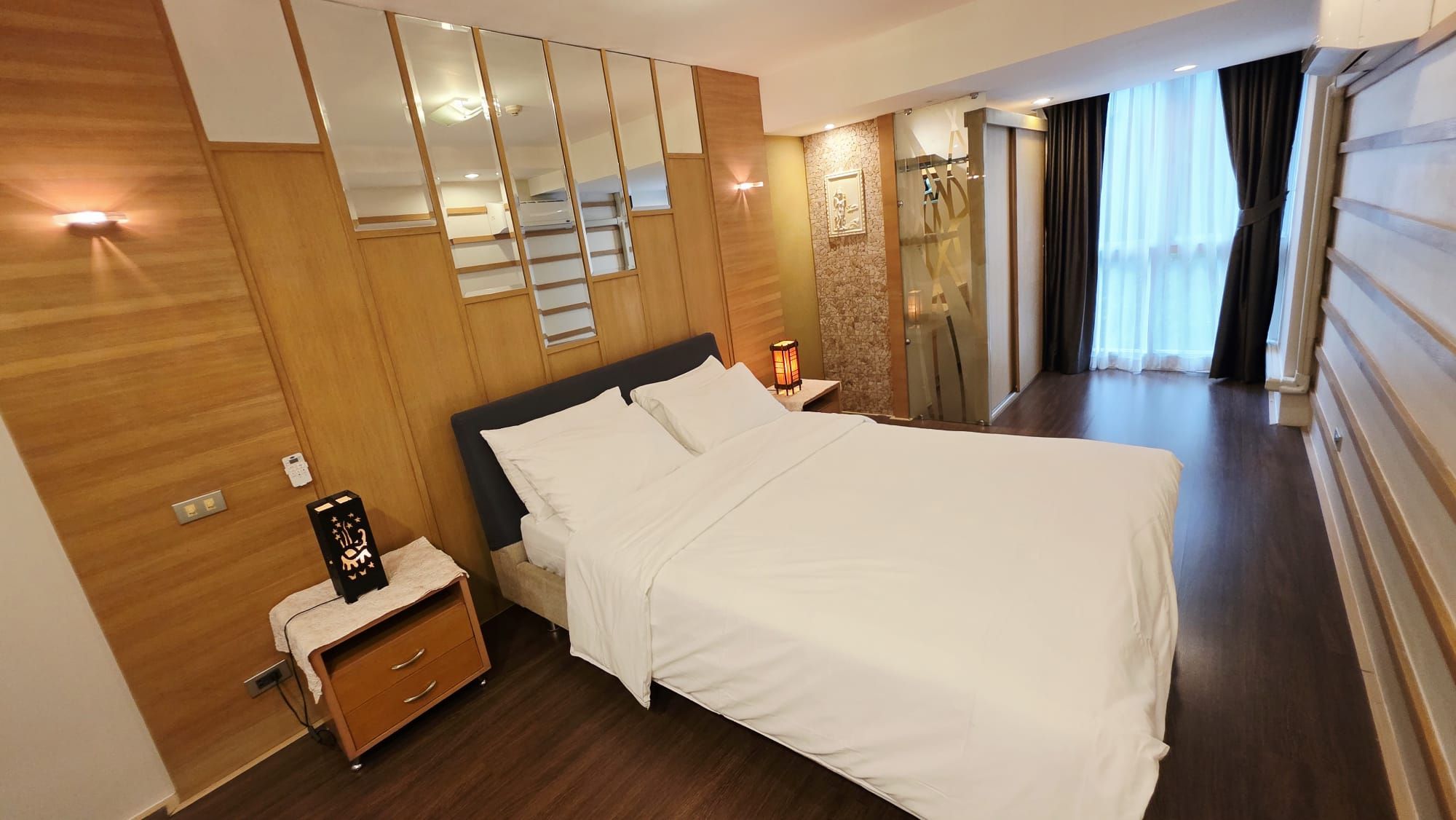 A hotel room with a large bed, wooden paneling, a nightstand, a lamp, a mirror, and a view of the outside through a window.
