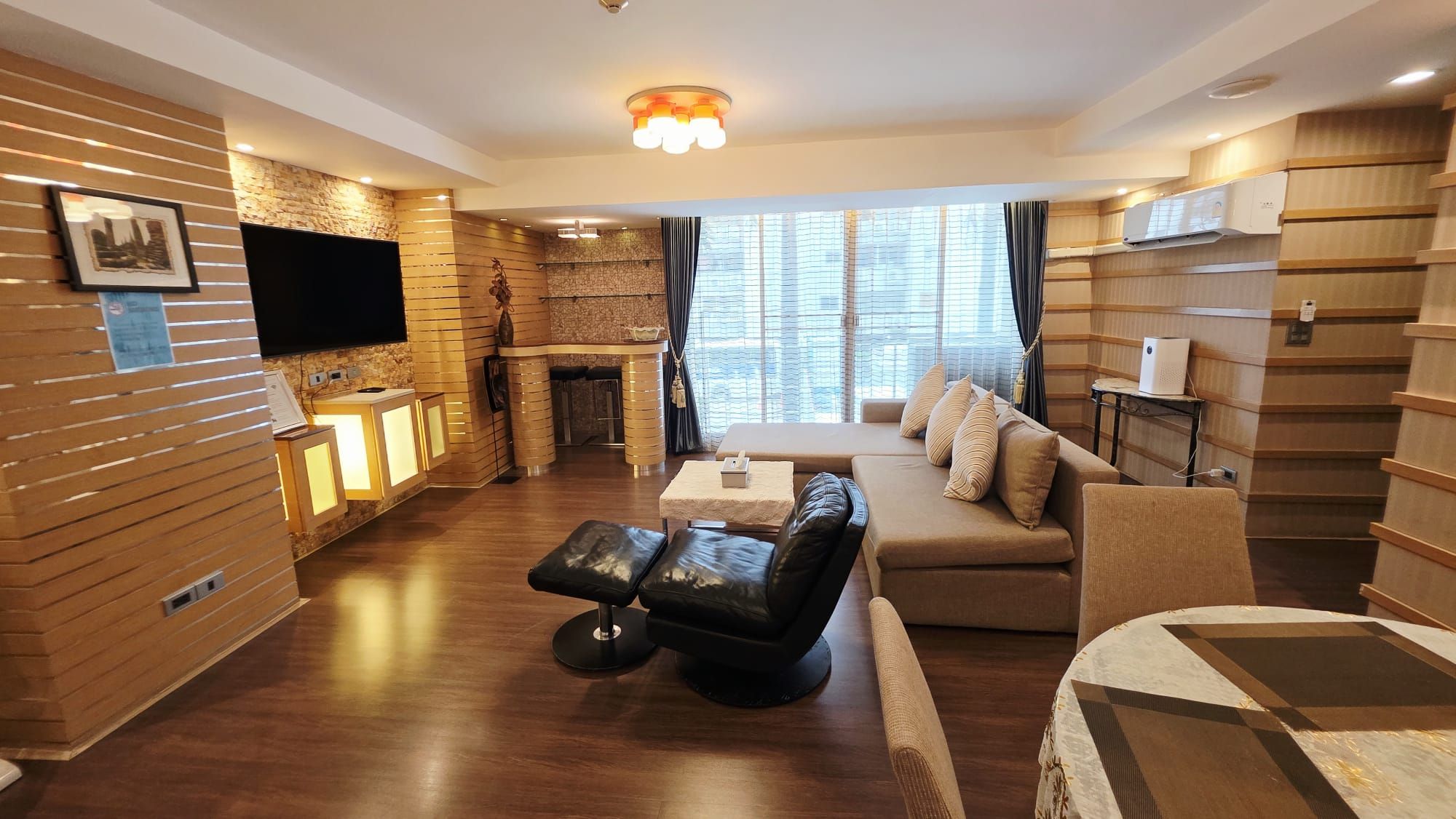 Ein modernes Wohnzimmer mit einer Couch, einem schwarzen Ledersitzhocker, einem kleinen Tisch, einem Kamin und einem an der Wand befestigten Fernseher.