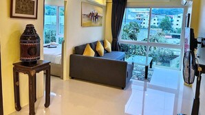 A living room with a sofa, a table with a decorative item, a painting, and a large window with a view of buildings. - 6-35 - 3 Bdr Condo with Balcony and shared Pool (Tambon Patong)