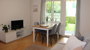 Ein modernes Wohnzimmer mit Fernseher auf einer weißen Kommode, einem Esstisch mit Stühlen und einem Sofa.