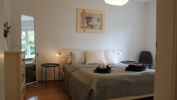 Ein Schlafzimmer mit Bett, Nachttischen, einem Sessel, einem Schreibtisch, einem Spiegel und einer Pendelleuchte.