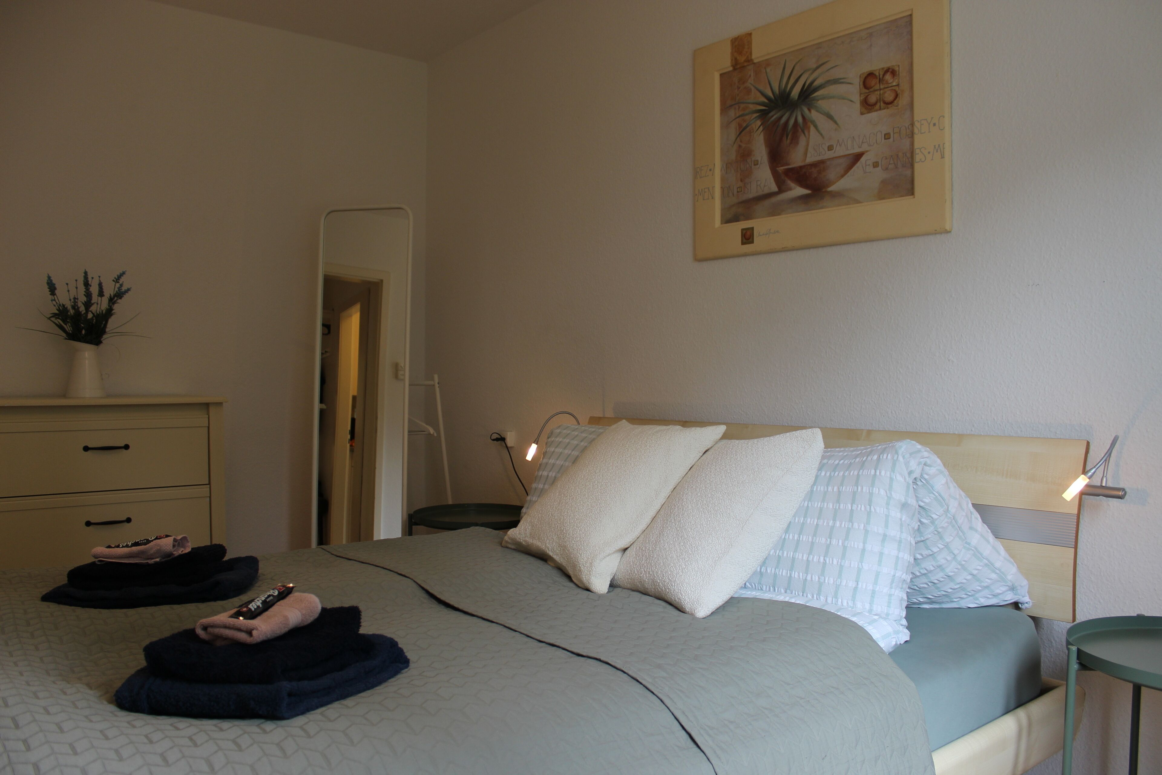 A neatly made bed with pillows and folded towels, a bedside table with a lamp, and a wall-mounted painting.