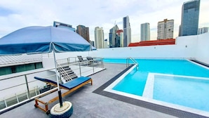 Una piscina en la azotea con un horizonte urbano de fondo, un área techada con sillones de descanso y una escalera que conduce a un nivel superior.