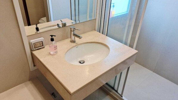 A bathroom with a beige sink, a mirror, a handheld soap dispenser, and a window with blinds.