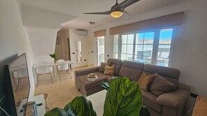 Una sala de estar con sofá, una mesa de comedor, sillas, ventilador de techo y ventanas grandes con vistas.