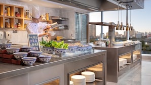 Een moderne keuken met een kok die voedsel bereidt, diverse kommen en borden op het aanrecht, en een stadszicht door grote ramen.