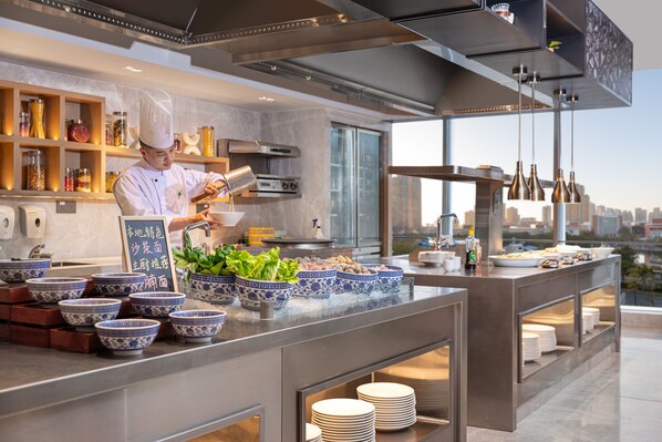 A modern kitchen with a chef preparing food, a variety of bowls and plates on the counter, and a city view through large windows. - Holiday Inn Quanzhou Donghai by IHG (Quanzhou)