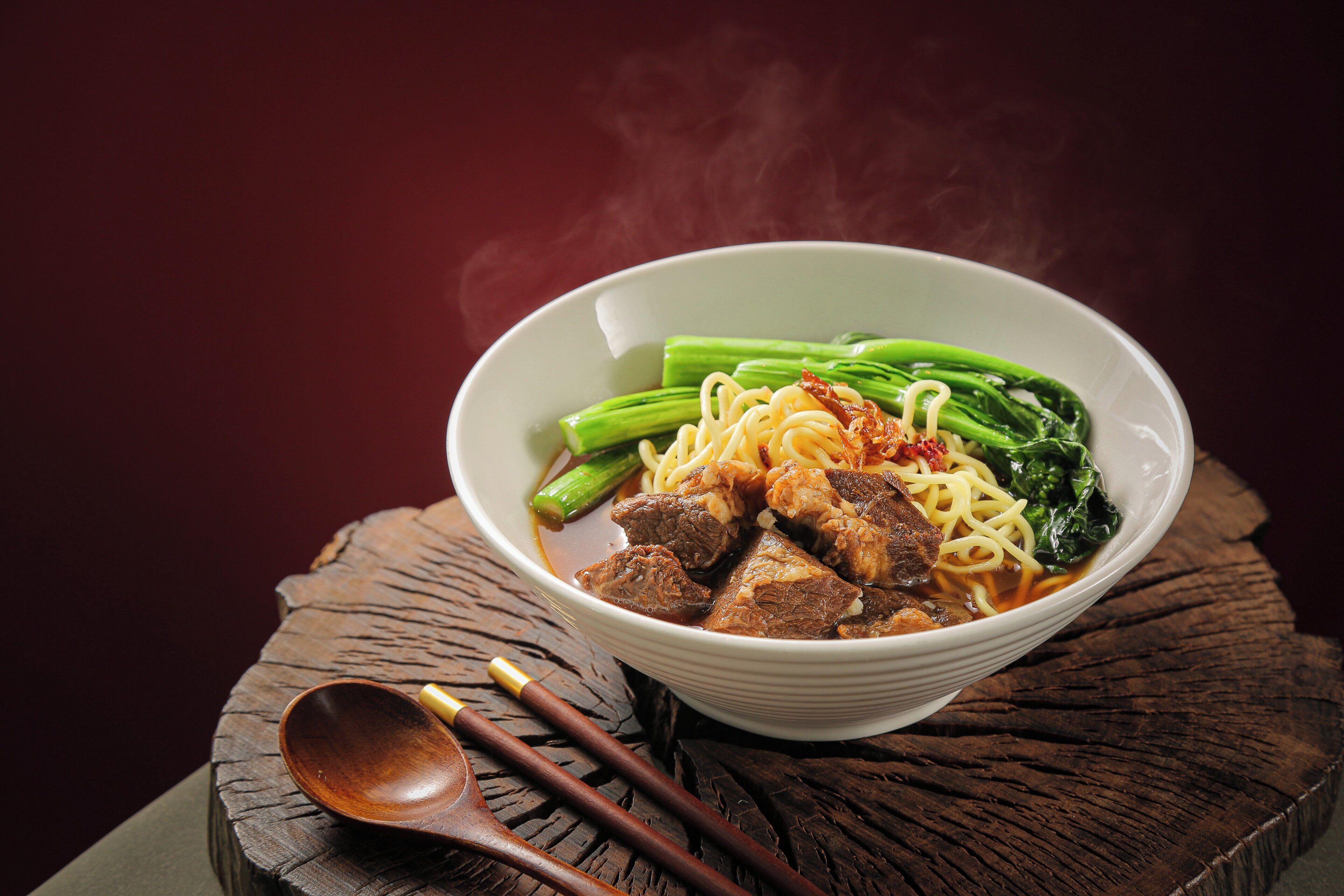 A bowl of noodles with meat and vegetables on a wooden surface.
