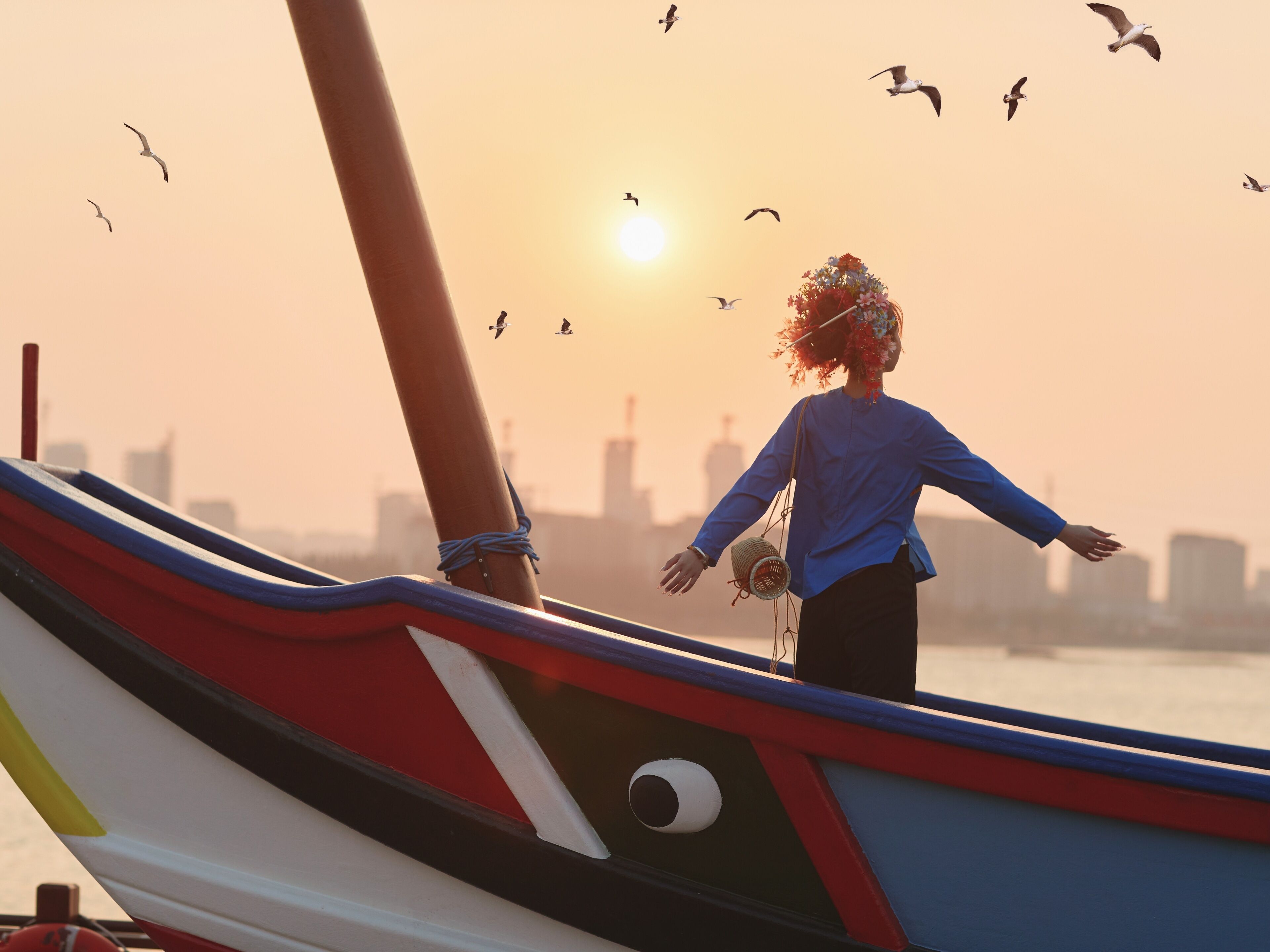 Una persona de pie en un bote, con los brazos extendidos, mirando al atardecer con aves volando al fondo.