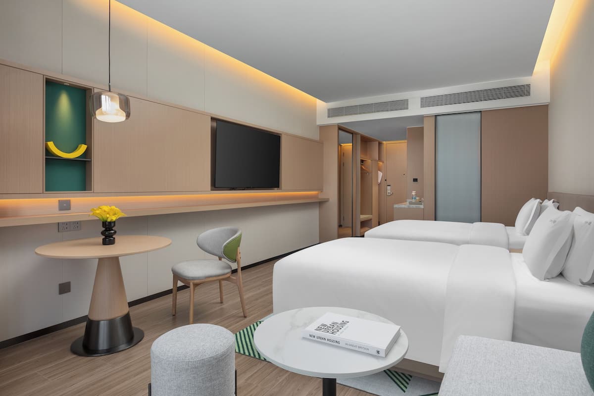 Una habitación de hotel moderna con una cama grande, una pequeña mesa redonda con un libro, una silla, un televisor montado en la pared y una mesita con un jarrón de flores.