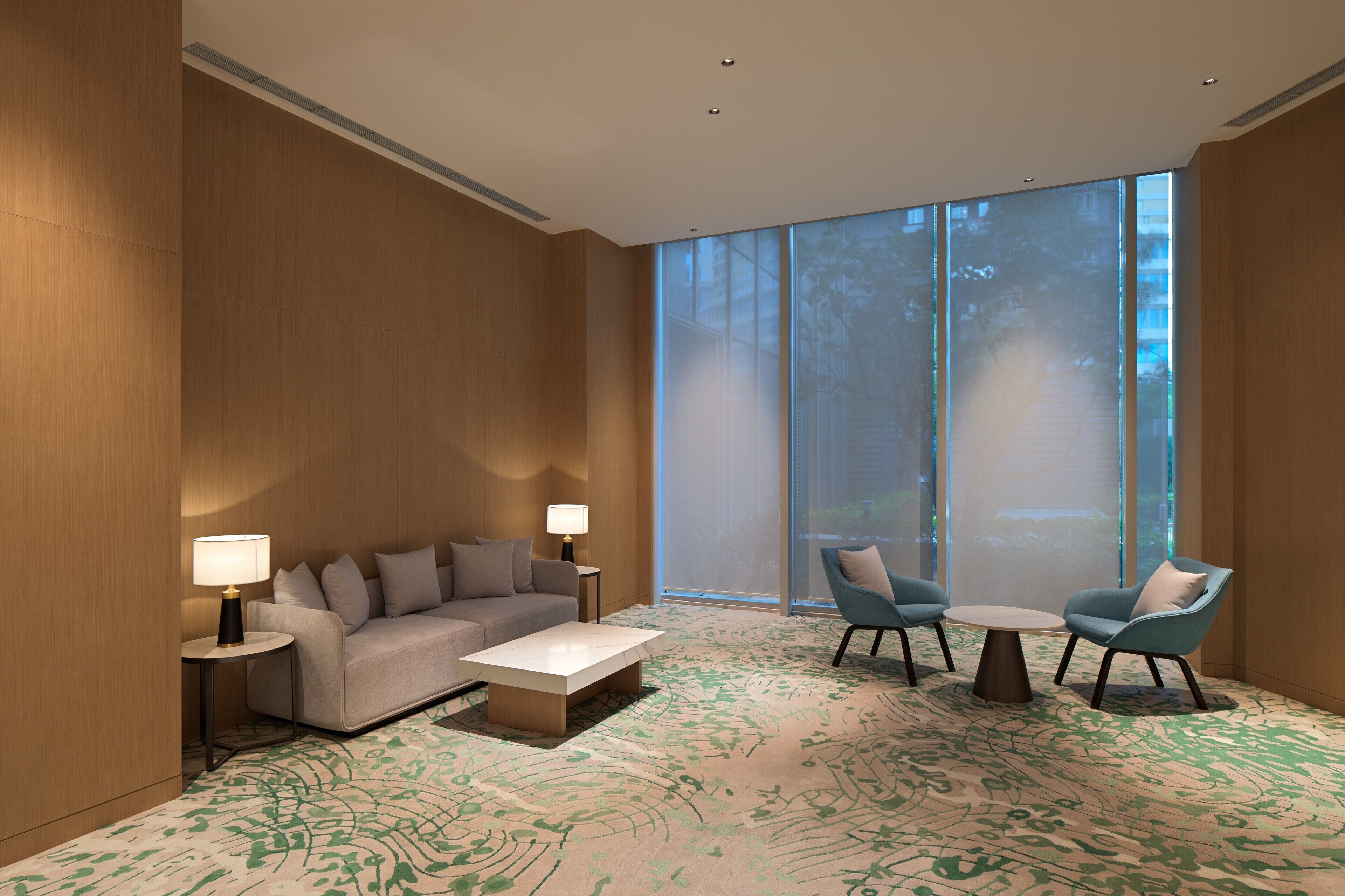 A modern hotel room with a large window, a sofa, two armchairs, a coffee table, and two lamps.