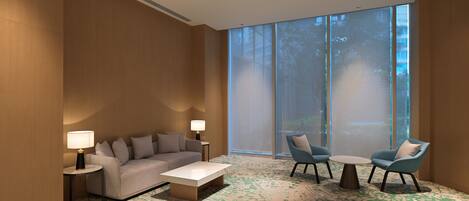 A modern hotel room with a large window, a sofa, two armchairs, a coffee table, and two lamps.